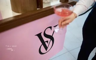 Victoria's Secret event bar with the VS logo on a pink panel and a hand holding a cocktail